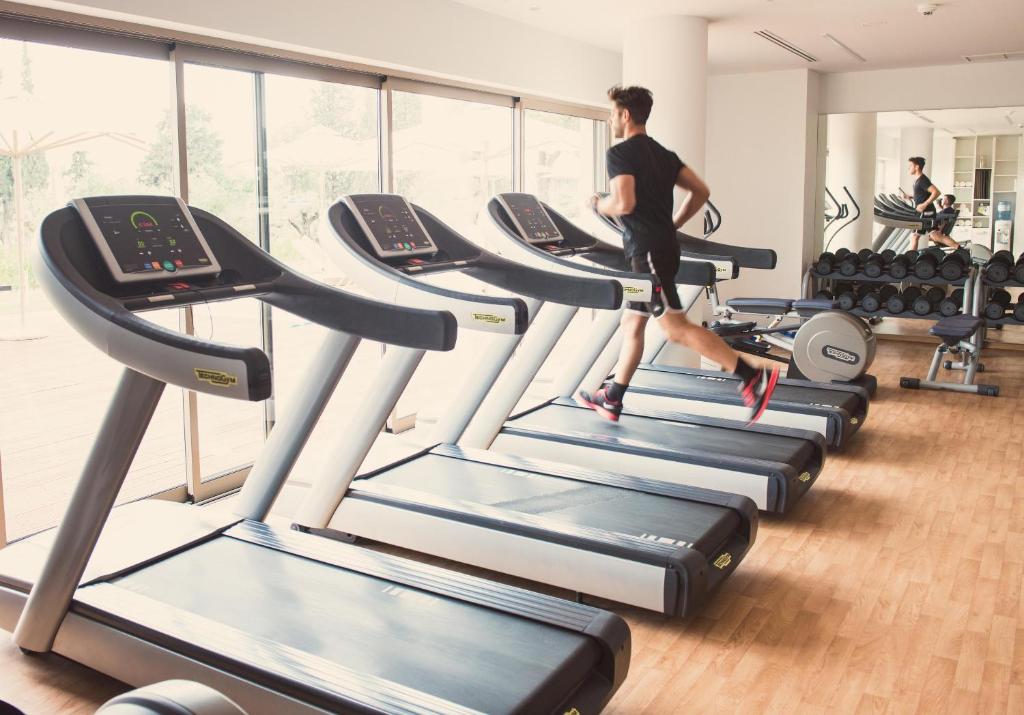 Vīrietis skrien uz skrejceliņa modernā sporta zālē, kur redzami vairākas skrejceliņu ierīces un svaru trenažieri. Gaisma ir gaiša un telpa ir plaša.
