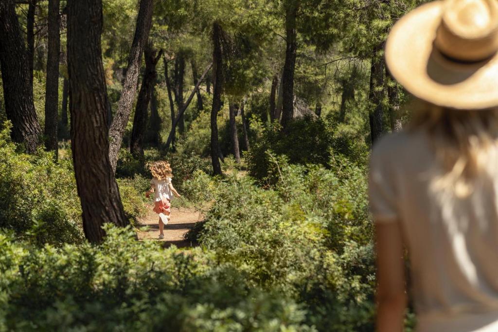 Meitene skriež pa meža taku, kamēr pieaugušais stāv fonā, baudot dabu. Ainava ir zaļa un saulaina, ar augstiem kokiem un krūmiem.