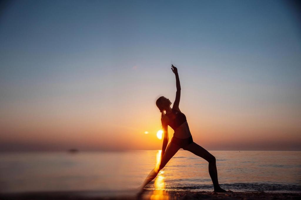 Sieviete veic jogas pozas pludmalē saulrieta laikā, kad debesis ir krāšņas ar siltām krāsām.