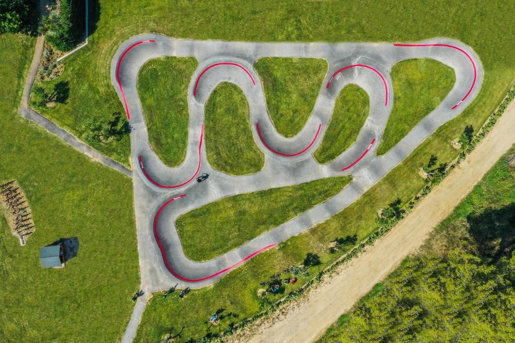 Asfaltēta trase ar sarkanām malām, kas veido sarežģītu līkumu modeli, atrodas zaļā pļavā. Attēlā redzams arī cilvēku grupējums un tuvumā esoša māja.