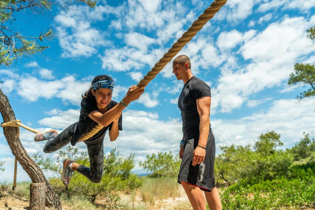 Bērns izpilda akrobātiskus trikus, šūpojoties no virves zem saulainām debesīm, kamēr pieaugušais skatās un sniedz atbalstu. Skats uz dabu fona.