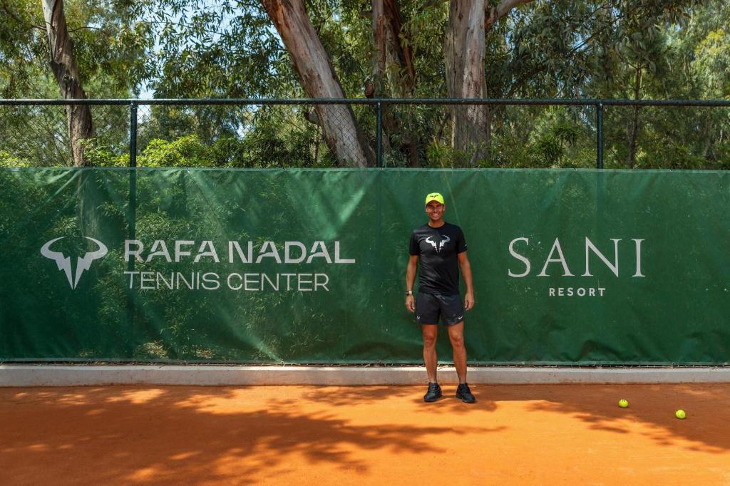 Rafa Nadal tenisa centra reklāma ar zaļu fona baneri, uz kura redzami centrs nosaukums un Sani kūrorta logo. Attēlā redzams vīrietis sportiskā apģērbā, kas stāv uz tenisa laukuma.