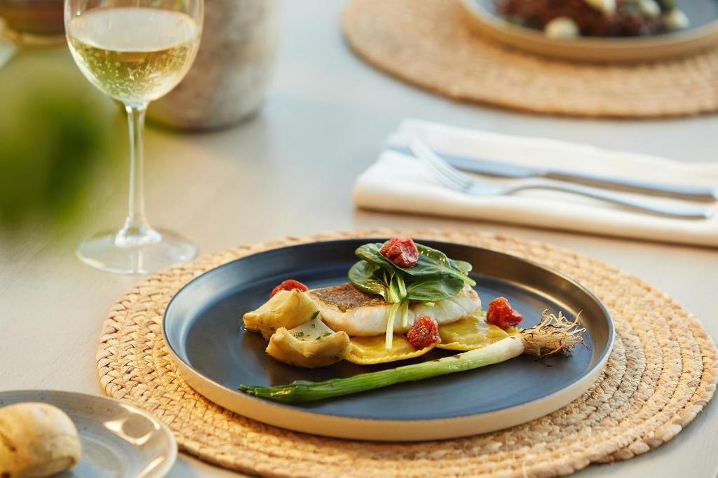 Eleganta maltīte ar ceptu zivi, dekorēta ar dārzeņiem un garšaugiem, pasniegta uz melnas šķīvja, blakus ir glāze baltā vīna.