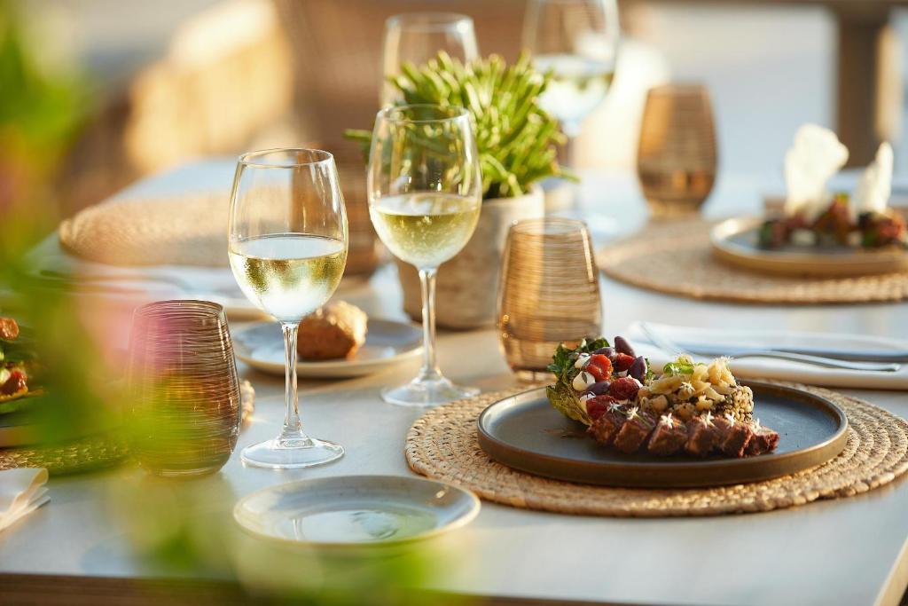 Elegants galda klājums ar baltvīnu un izsmalcinātām ēdienu porcijām, radot patīkamu un viesmīlīgu atmosfēru. Daba un gaisma piešķir noskaņu.