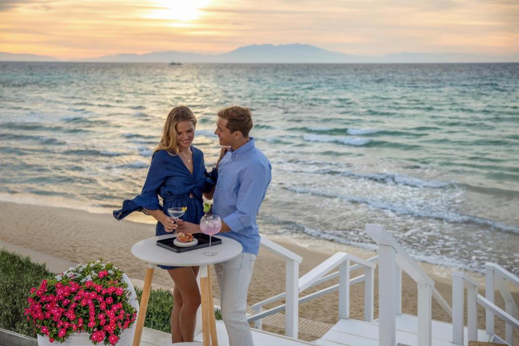 Romantisks brīdis pludmalē, kur pāris bauda dzērienus un uzkodas, fonā redzama saulrieta ainava.