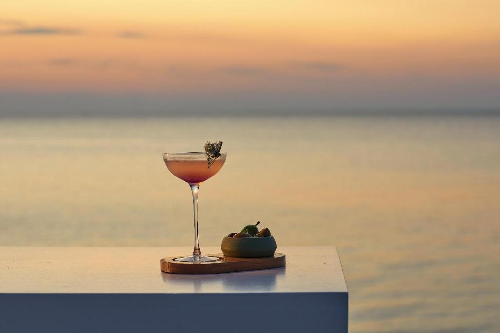 Elegants kokteilis ar garšaugu rotājumu, novietots uz galda pie saulrieta, fona veido mierīga jūra. Blakus atrodas maza trauciņš ar olīvām.