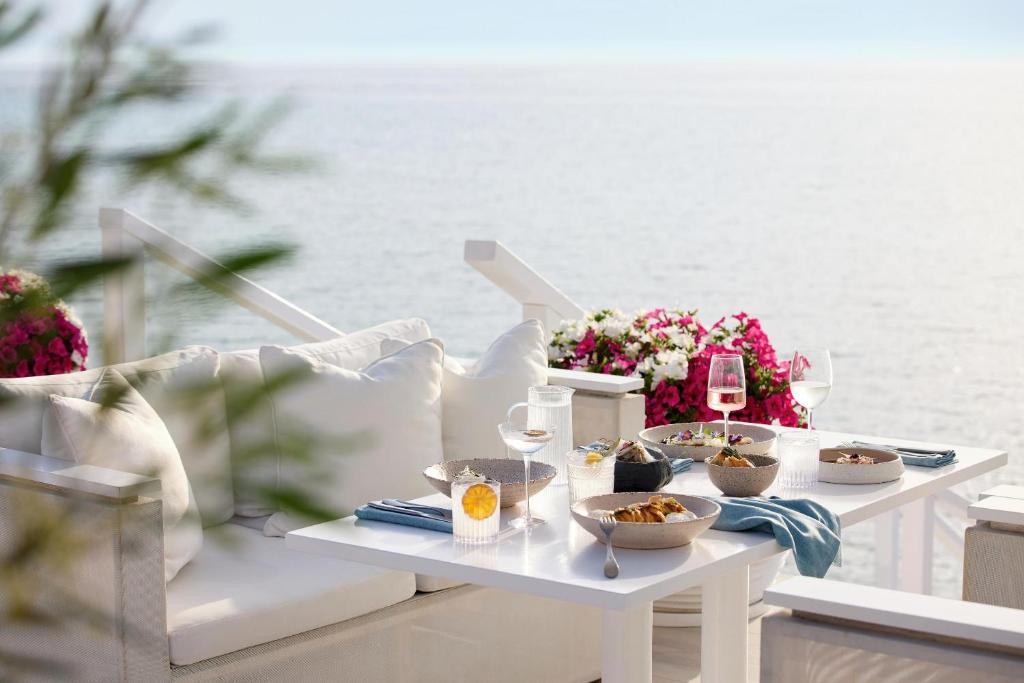 Elegantā maltīte uz terases ar skatu uz jūru, kurā ir dažādi ēdieni un dzērieni, ieskaitot vīnu un desertus. Pavasara ziedi pie galda papildina atmosfēru.