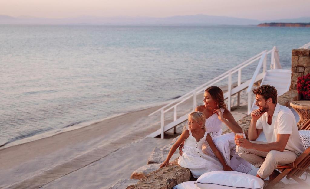 Ģimene, kas relaksējas pludmalē, skatoties uz saulrietu virs jūras, ģērbtas baltos apģērbos. Siltas krāsas un mierīga atmosfēra rada patīkamu brīvdienu sajūtu.