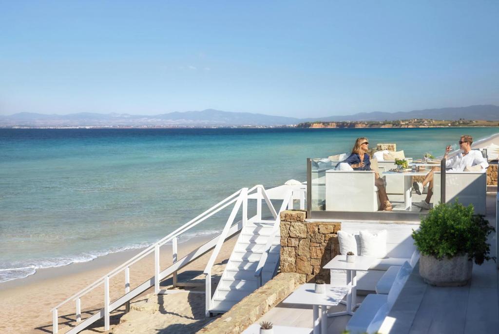 Divi cilvēki relaksējas pie jūras, baudot sauli un mierīgu atmosfēru, sēžot modernā terasē ar skatu uz pludmali un zilo ūdeni.