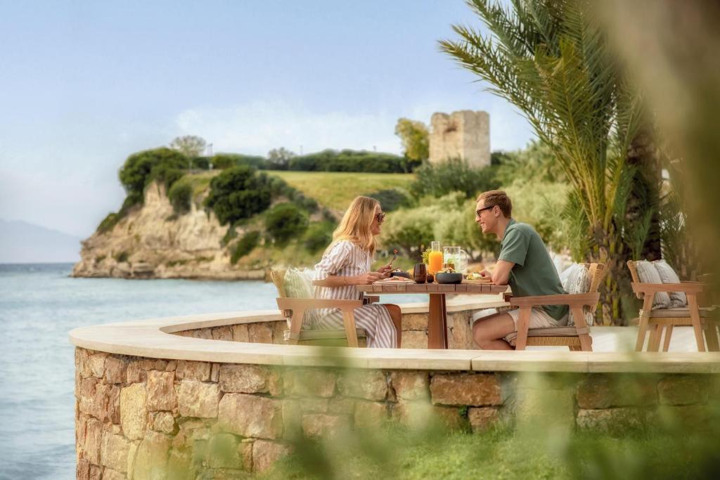 Romantiska brokastis pie jūras, pāris bauda ēdienu un dzērienus skaistā ainavā ar kalniem un ūdeni fonā.