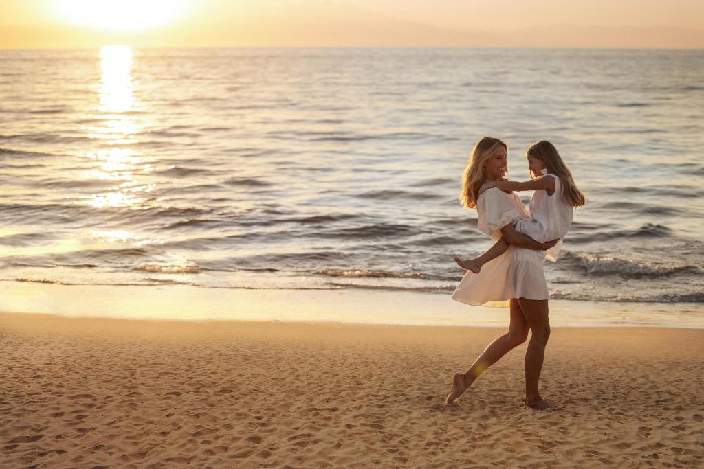 Māte un meita kopā priecīgi pastaigā pludmalē saulrietā, izbaudot mirkļa burvību un tuvību. Ūdens viļņi maigi skar smiltis, radot mierīgu atmosfēru.