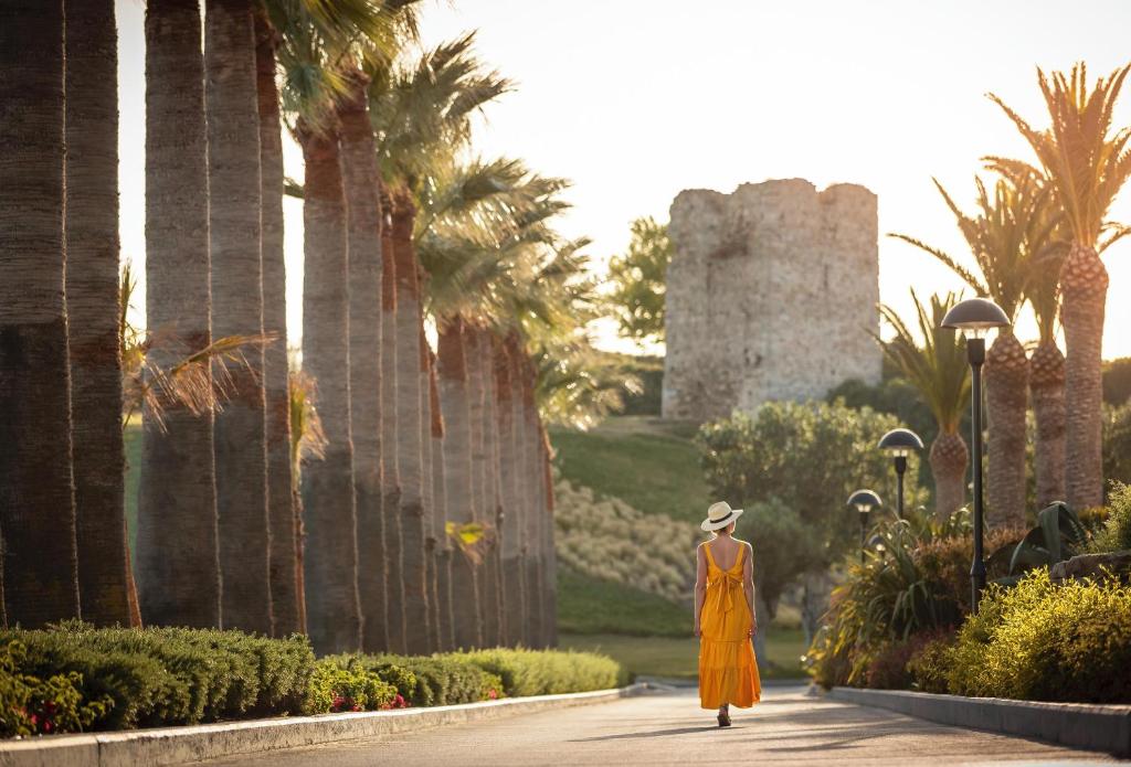 Sieviete dzeltenā kleitā pastaigājas gar palmām, saulē apgaismotā ainavā, fona atrodas veca mūra tornis.