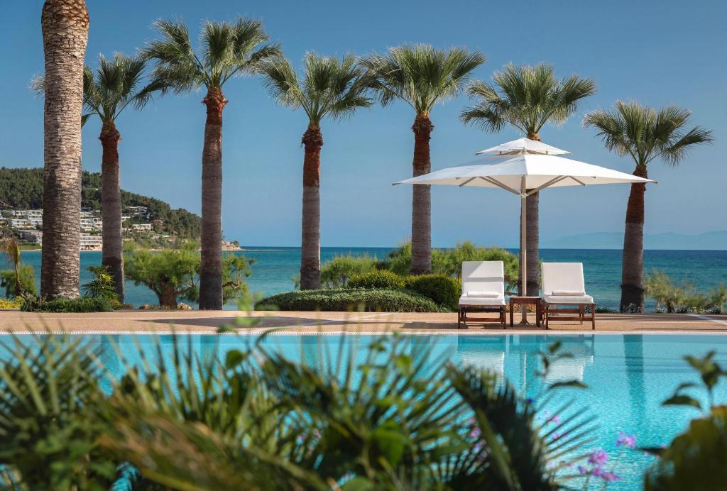 Viesmīlīgu pludmales skatu ar palmām un baseinu, kurā ir divi atpūtas krēsli zem balta sauleslaika. Jūras viļņi un kalnu ainava veido relaksējošu atmosfēru.