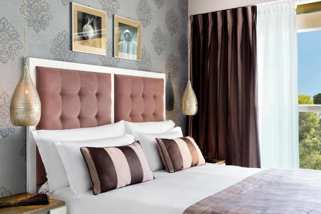 Mūsdienīgs guļamistabas interjers ar rozā krāsas gultas galvu, dekoratīvām spilveniem un elegantiem apgaismojuma elementiem. Sienas rotā stilizēti attēli, pie loga ir tumši un gaisīgi aizkari.