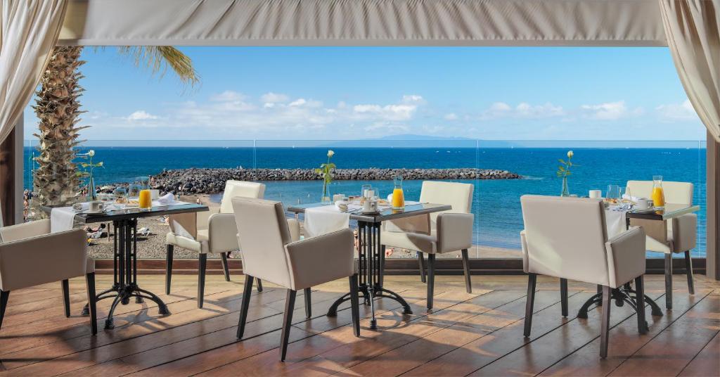 Restorāna interjers ar skatu uz jūru, kur baltas galds un krēsli ir izvietoti pie plaša loga, nodrošinot brīnišķīgu ainavu ar pludmali un zilu ūdeni.
