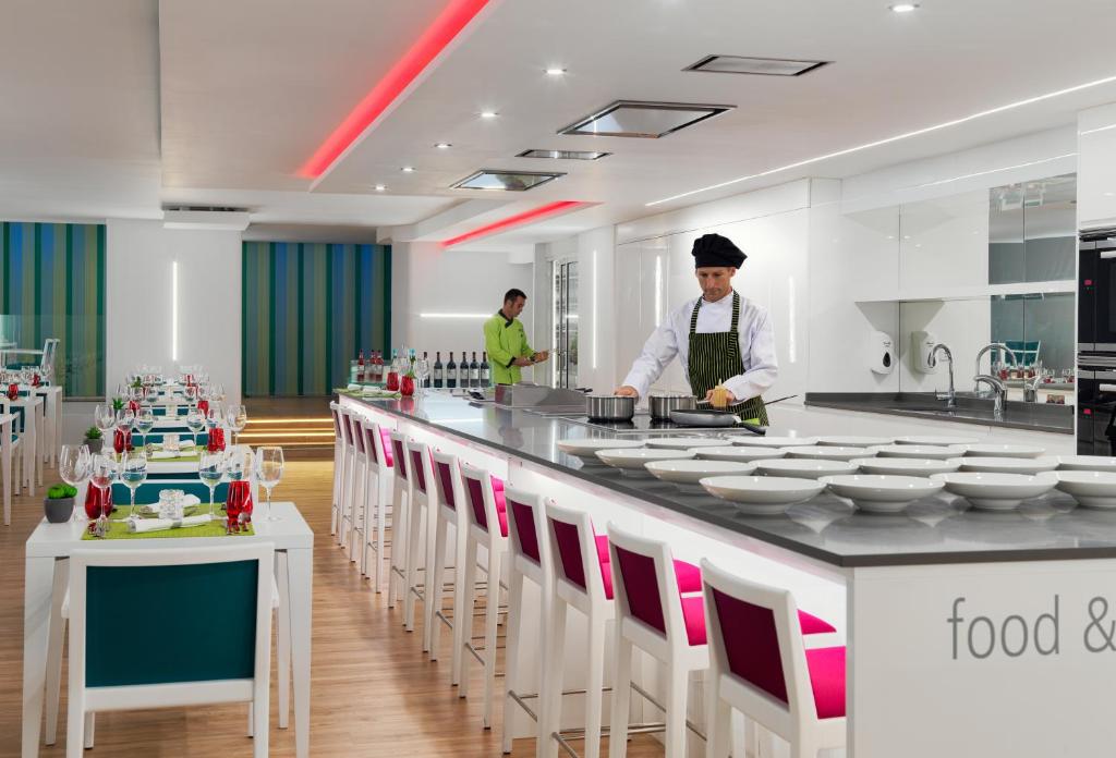 Mūsdienīga virtuve ar elegantiem galdiem un krāšņu apgaismojumu, kur šefpavārs gatavo ēdienus. Interjerā ir gaišas krāsas un stilīgi krēsli, radot viesmīlīgu atmosfēru.