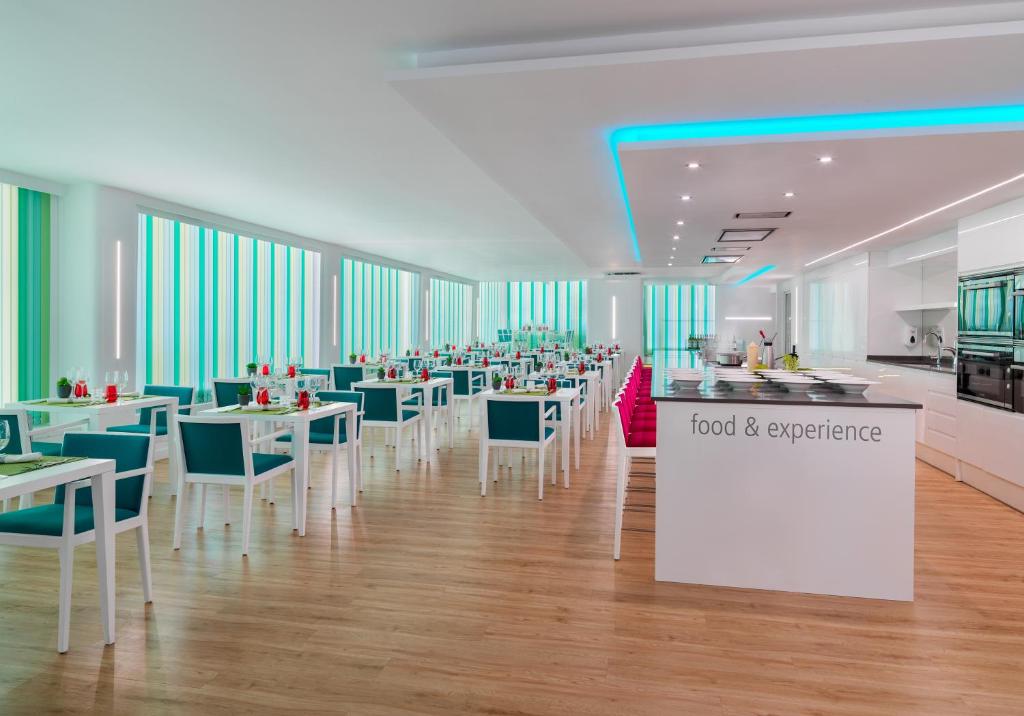 Mūsdienīgs restorāns ar gaišu interjeru un stilīgām krēsliem, izceļot krāsainus logus un elegantu galdu uzstādījumu. Ideāla vieta gastronomiskām pieredzēm.