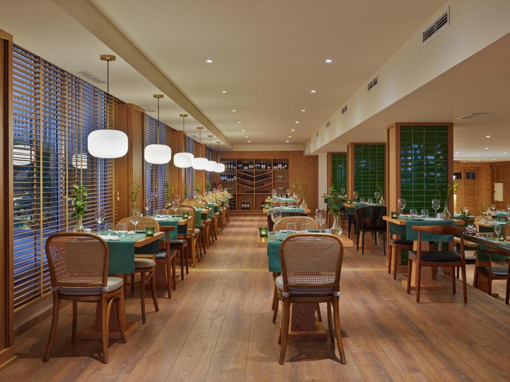 Mūsdienīgs restorāns ar koka interjeru, zaļu galdu segumu un gaišām lustām, radot relaksējošu atmosfēru. Logi ar žalūzijām nodrošina dabīgu apgaismojumu un skatu uz ārpusi.