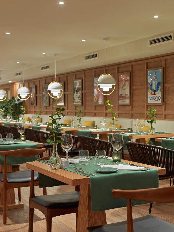 Mūsdienīgs restorāna interjers ar elegantiem galdiem, zaļiem galdautiem un dekoratīvām augu kompozīcijām. Sienas rotā vintage plakāti, radot patīkamu atmosfēru.