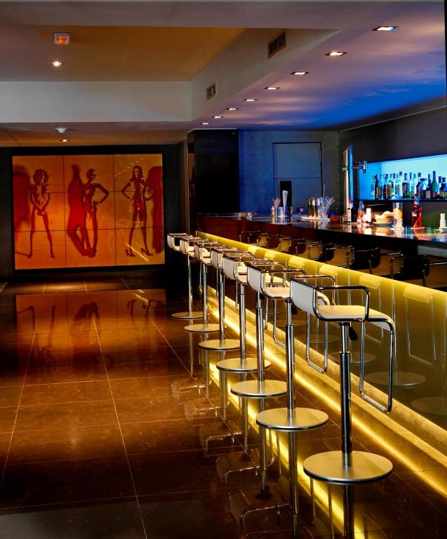 Mūsdienīgs bārs ar stilīgām krēslām un izsmalcinātu apgaismojumu, kas rada patīkamu atmosfēru. Sienā redzama krāsaina mākslas gleznas kompozīcija.