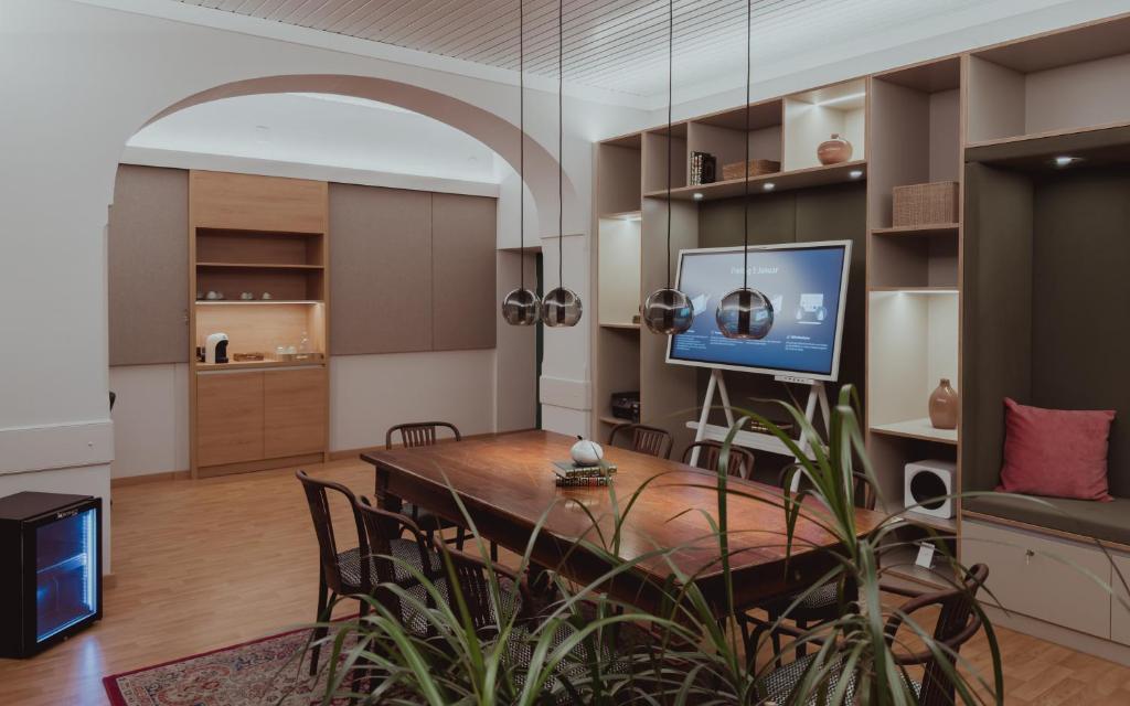 Mūsdienīga sapulču telpa ar koka galdu, stilīgām lampām un televizoru, kas piedāvā prezentācijas iespējas. Telpā ir dekoratīvi elementi un augu klātbūtne, radot patīkamu atmosfēru.