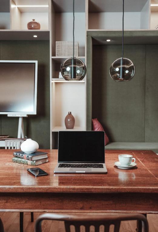 Mūsdienīgs darba galds ar klēpjdatoru, grāmatām un kafijas krūzi, apgaismots ar stilīgām laternām. Fons ar televizoru un dekoratīviem elementiem rada mājīgu atmosfēru.
