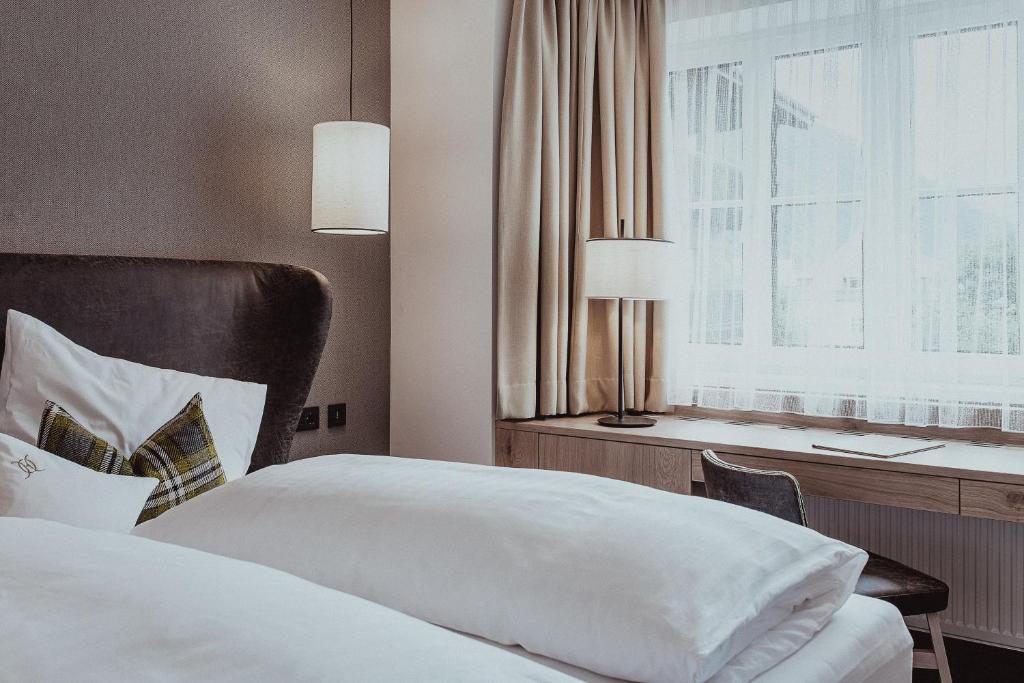 Mūsdienīgs guļamistabas interjers ar ērtu gultu, mīkstiem spilveniem un dabīgu apgaismojumu. Loga tuvumā ir rakstāmgalds un stilīga lampiņa, kas rada mājīgu atmosfēru.