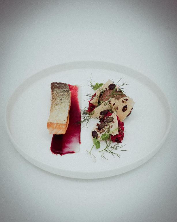 Grezns ēdiens, kurā redzama cepta zivs fileja ar kraukšķīgu ādu un sarkans mērces pleķis, blakus ir dekorēts salātu ēdiens ar zaļumiem un riekstiem.