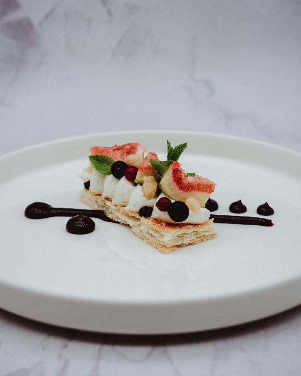 Deserts ar kārtainās mīklas bāzi, apdarināts ar krēmīgu pildījumu, ogām un svaigiem augļiem. Uz šķīvja ir dekoratīva šokolādes mērce.