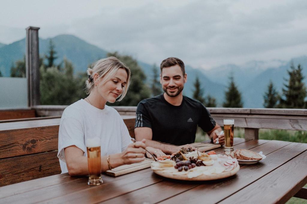 Divi cilvēki izbauda maltīti ārā uz terases, ar kalnu ainavu fonā. Uz galda ir pasniegti dažādi uzkodas, un katram ir dzēriens.