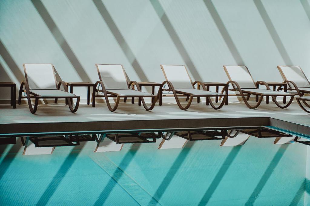 Balto krēslu rinda pie baseina ar atspīdumu ūdenī, radot mierīgu un relaksējošu atmosfēru. Gaismas un ēnu spēles uz sienas piešķir telpai estētisku pievilcību.