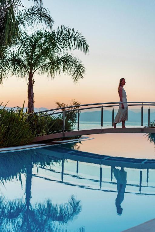 Sieviete, kas staigā pāri tiltam virs baseina, apņemta ar palmām un saulrieta gaismu, radot mierīgu un relaksējošu atmosfēru.