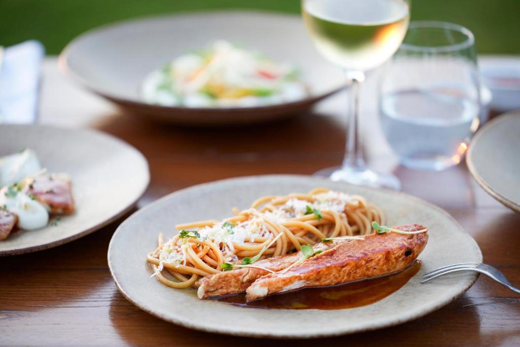 Svaigi pagatavota pastas porcija ar laša fileju, pasniegta uz elegantas plāksnes, blakus glāzei baltvīna un ūdens. Fons papildina maltīti ar citiem ēdieniem.