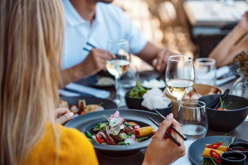 Vasaras maltīte ar svaigiem dārzeņiem un vīnu, cilvēki izbauda gardus ēdienus. Galda klājumā redzami dažādi trauki un virtuves piederumi.