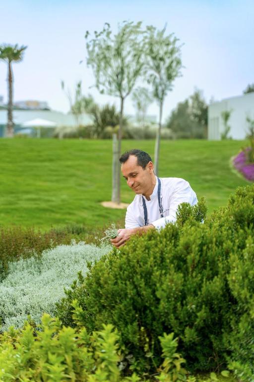 Vīrietis dārzā rūpējas par augiem, pievēršoties zaļajai videi un dārzkopībai. Fons ar zaļām zālēm un kokiem rada mierīgu atmosfēru.