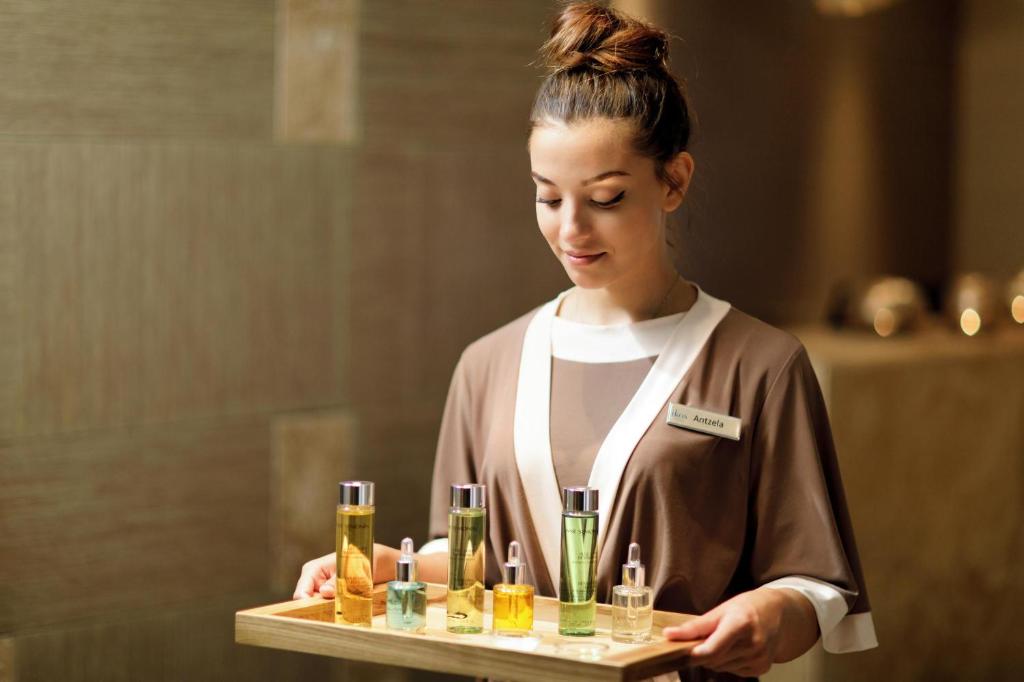 Skaistumkopšanas speciāliste, turot koka paplāti ar dažādiem ādas kopšanas produktiem, piemēram, eļļām un serumiem. Fons ir elegants un modernais interjers.