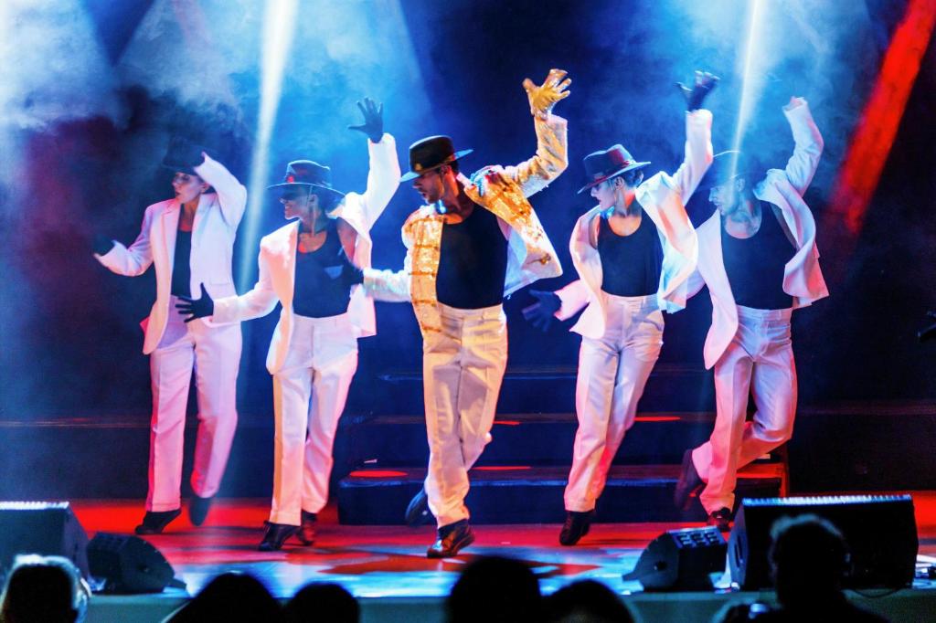 Deju izpildītāji uz skatuves, tērpušies baltos uzvalkos un melnās cepurēs, izpilda sinhronas kustības, kamēr apgaismojums un dūmi rada dinamisku noskaņu.