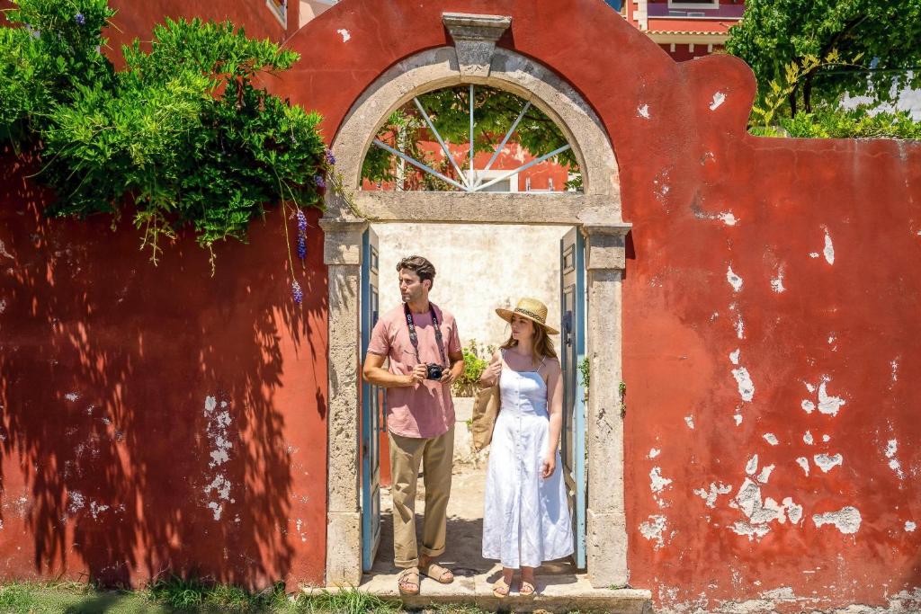 Pāri sarkaniem mūriem izveidots arku formas vārtiņš, caur kuriem divi jaunieši pozē saulainā dienā. Vīrietis tur kameru, bet sieviete valkā baltu kleitu un platvērti.