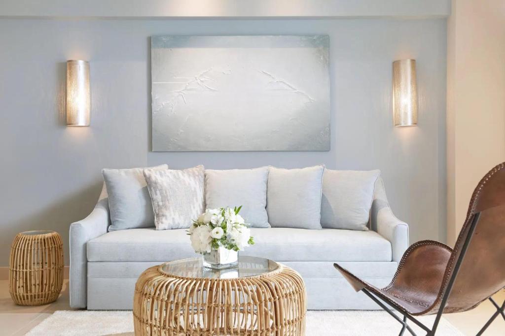 Mūsdienīgs viesistabas interjers ar pelēku dīvānu, rattan galdiņu un ziedu kompozīciju. Sienas apgaismojums un mākslas darbs pie sienas piešķir telpai elegantu atmosfēru.