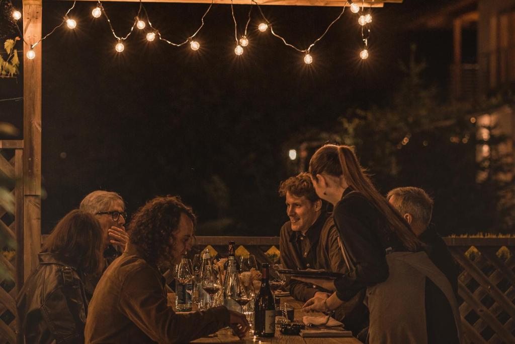 Vakara gaismas apgaismotā terasē grupa cilvēku bauda vakariņas, sarunājoties un smejoties. Galda virsma ir klāta ar vīna pudelēm un glāzēm, radot siltu un draudzīgu atmosfēru.