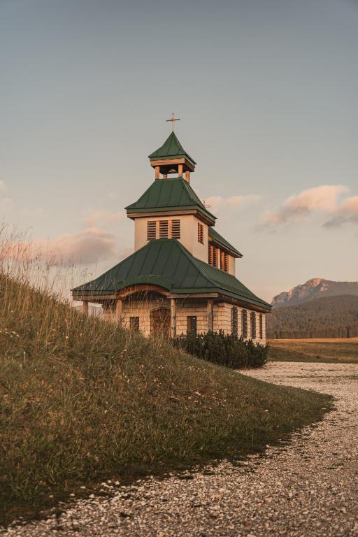 Vēsturiska koka baznīca ar zaļu jumtu, ieskauta dabā, ar kalniem fonā. Šis attēls parāda mierīgu ainavu un arhitektūras skaistumu.