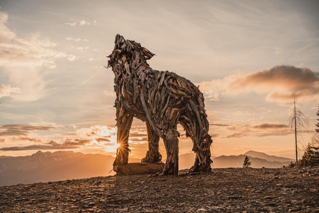 Koka skulptūra, kas attēlo vilku, stāv kalnu ainavā saulrieta gaismā, radot iespaidīgu un māksliniecisku atmosfēru.