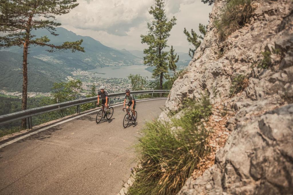 Divi riteņbraucēji brauc pa kalnu ceļu, ar gleznainu skatu uz ezeru un kalniem fona. Ainava ir zaļa un kalnaina, radot ideālu vidi aktīvai atpūtai.