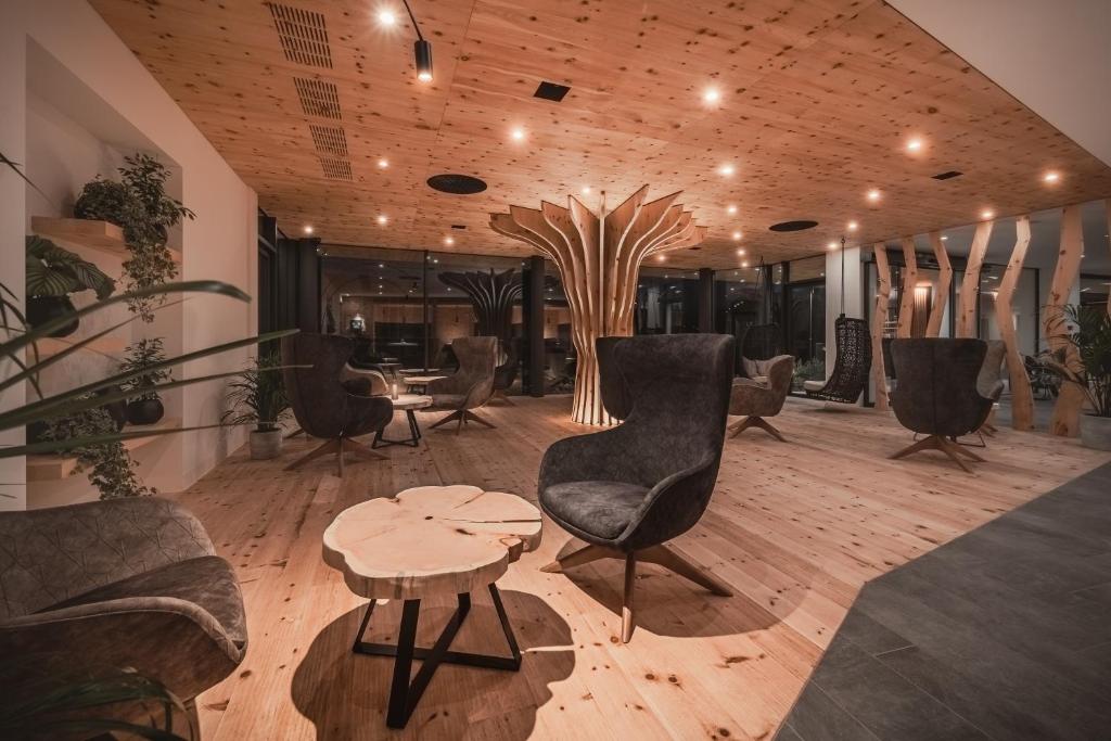 Mūsdienīgs interjers ar koka grīdu un elegantiem krēsliem, centrā izceļas stilizēts koka elements. Telpa ir apgaismota ar siltām gaismām, piešķirot mājīgu atmosfēru.