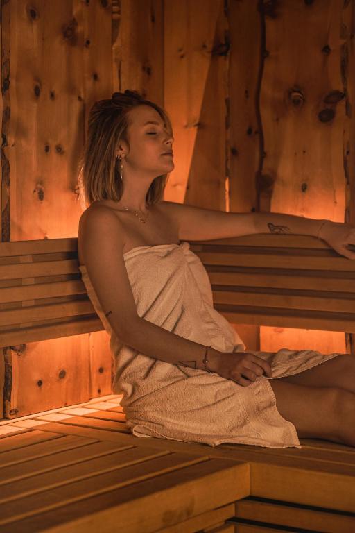 Jaunā sieviete atpūšas koka pirtī, apņemta ar dvieli un baudot siltumu. Apkārt ir koka sienas ar maigu apgaismojumu, kas rada relaksējošu atmosfēru.