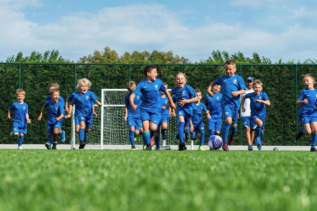Bērni skrien futbolā, tērpušies zilās formās, ar prieku un enerģiju laukuma vidū. Fons ietver futbola vārtus un zāles laukumu.