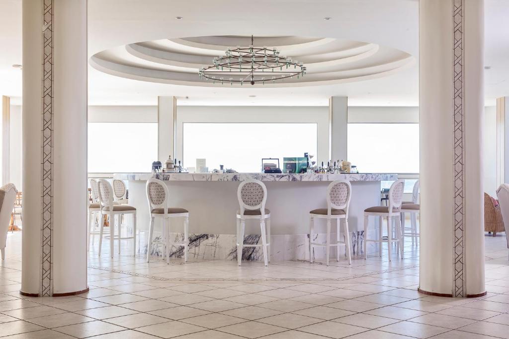 Mūsdienīgs bārs ar marmora virsmu, apgaismots ar elegantu lustru un stilīgām baltām krēsliem, radot relaksējošu un modernu atmosfēru. Plašas logu ailes nodrošina dabisko apgaismojumu un skatu uz ārpusi.