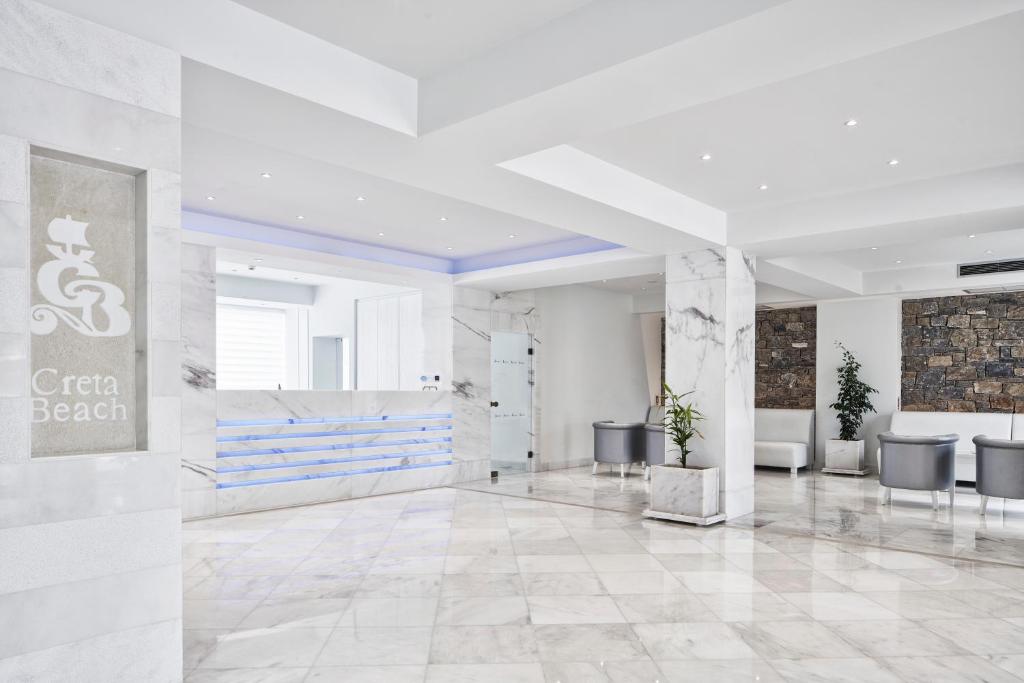Mūsdienīgs viesnīcas reģistratūras interjers ar marmora grīdu, gaišām sienām un modernu apgaismojumu. Redzams viesnīcas "Creta Beach" logo un eleganti sēdvietas.