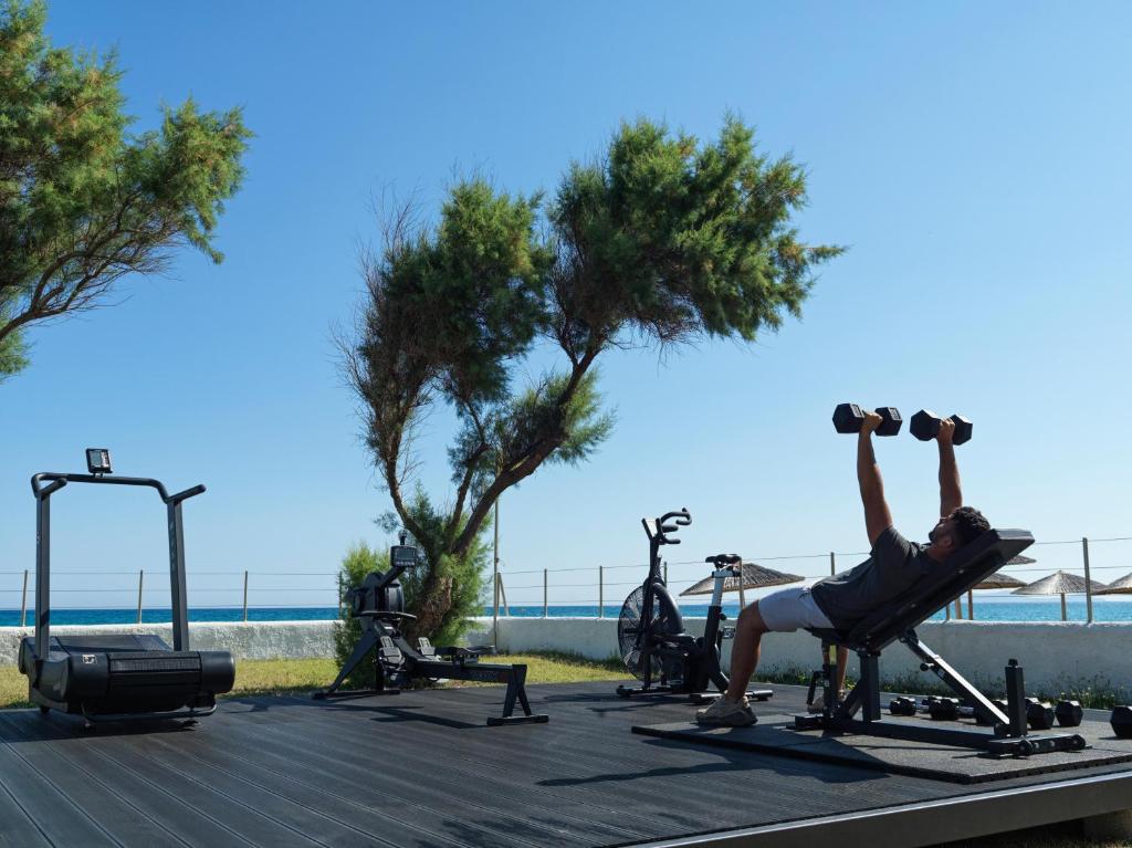 Treniņš brīvā dabā ar svaru stieni un kardio iekārtām, fons ar jūru un kokiem. Ideāla vide aktīvai atpūtai un fitnesa nodarbēm.