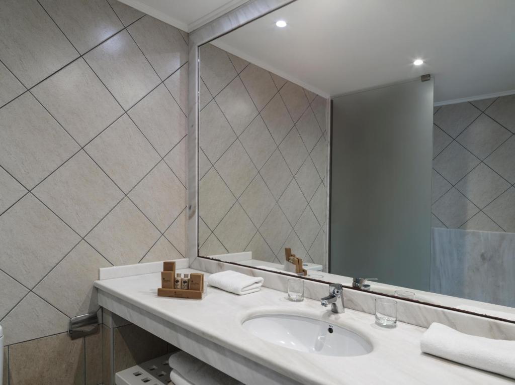 Moderns vannas istabas interjers ar flīžu sienām, diviem dvieļiem un higiēnas piederumiem uz izlietnes. Eleganta spoguļa un apgaismojuma kombinācija rada patīkamu atmosfēru.